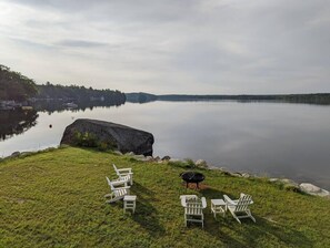 A lakeside view with a grassy area, several Adirondack chairs, and a black grill.