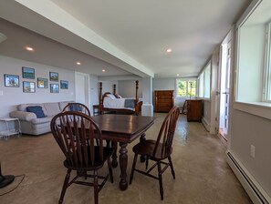 A spacious living room with a large dining table, a sofa, and a bed with a wooden headboard. There are framed pictures on the wall and a window providing natural light.