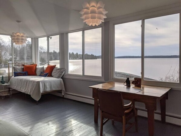 A room with a bed, a wooden table, and large windows overlooking a body of water.