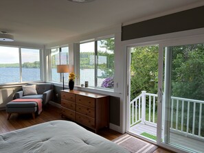 A bedroom with a large bed, a dresser, a chair, and a view of a lake through large windows. - Lakefront for 15, private beach and dock (Dedham)