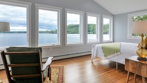 A bedroom with a large window overlooking a lake, a bed with a green throw, and a wooden chair. - Lakefront for 15, private beach and dock (Dedham)