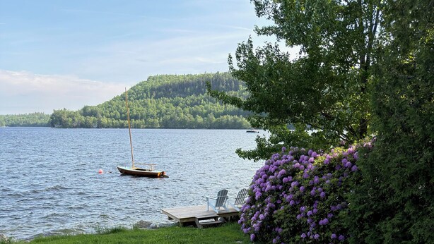 A wooden deck with a white railing overlooking a lake with a sailboat. - Lakefront for 15, private beach and dock (Dedham)