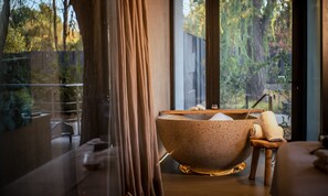 Zona de spa con una bañera independiente, un taburete de madera y una barandilla de vidrio con vistas a un jardín.