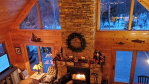 L’intérieur chaleureux d’un chalet avec une cheminée en pierre, des murs en bois et une grande fenêtre donnant sur un paysage enneigé.