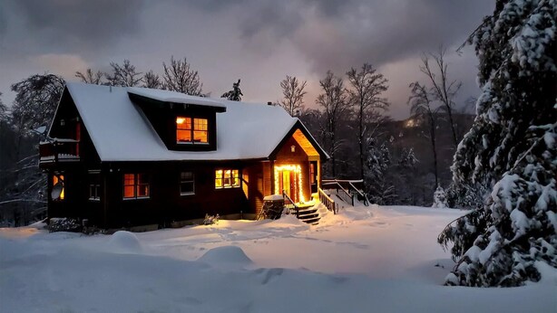 Une cabane en bois, dont le toit est recouvert de neige, avec une cheminée bien visible et une véranda dotée de marches menant à l’entrée.