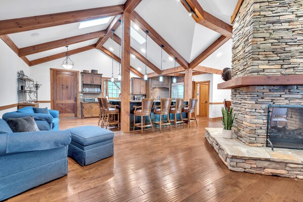 A spacious living area with a high ceiling, wooden beams, a stone fireplace, and a kitchen in the background.