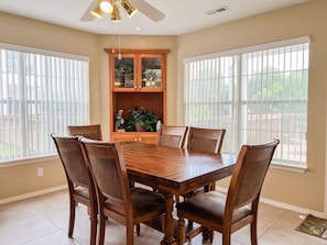 A dining room with a wooden table and chairs, a cabinet with glass doors, and large windows.