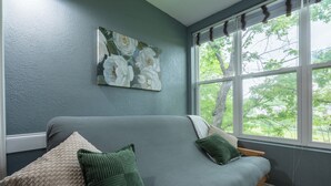 Ein Zimmer mit grauem Sofa, grünen Kissen, einem Blumenbild an der Wand und einem Fenster mit Blick auf Bäume.