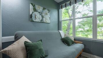 A room with a grey sofa, green cushions, a floral painting on the wall, and a window with a view of trees.