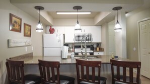 A modern kitchen with white cabinets, a stainless steel refrigerator, a built-in oven, and a granite countertop. There are three pendant lights, a welcome sign, and a door to the right.