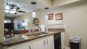A kitchen with granite countertops, a dishwasher, a clock, and a welcome sign.