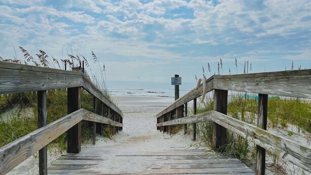 Un paseo marítimo de madera que lleva a la playa, con arena y césped a ambos lados.