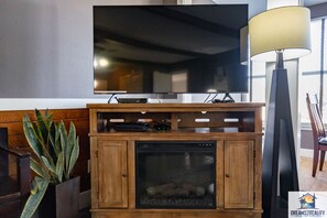 A living room with a flat-screen TV mounted on the wall, a fireplace with a glass door, a potted plant, and a floor lamp.