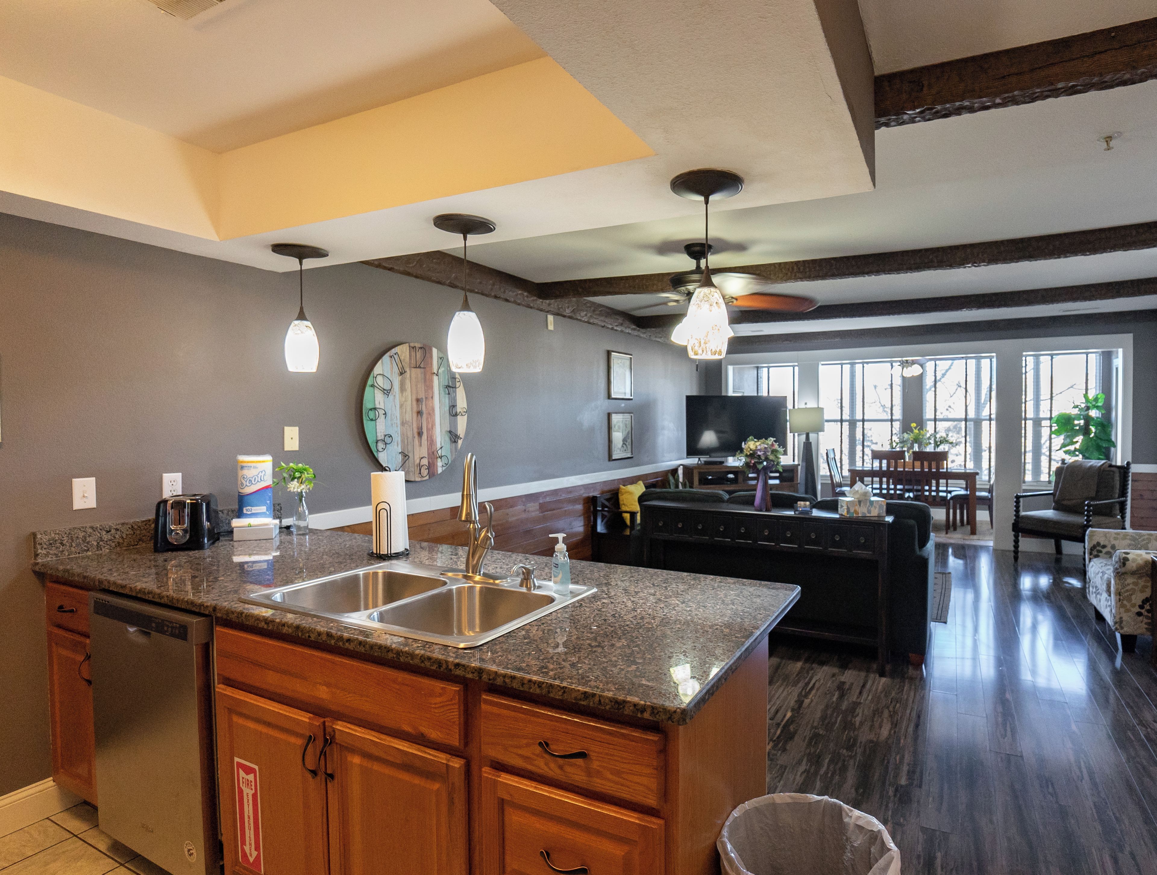 A spacious kitchen with granite countertops, a dishwasher, and a large island with a sink. There is a dining area with a sofa and a TV in the background.