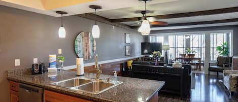 A spacious kitchen with granite countertops, a dishwasher, and a large island with a sink. There is a dining area with a sofa and a TV in the background.
