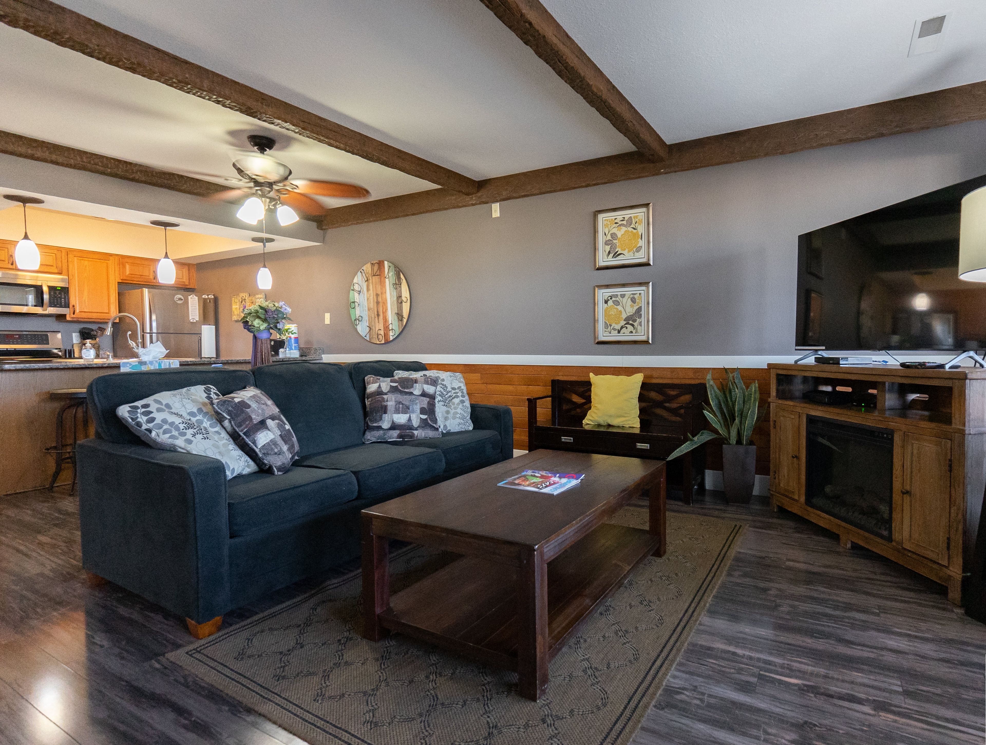 A living room with a sofa, coffee table, and a TV. There is a kitchen area with wooden cabinets and a refrigerator.
