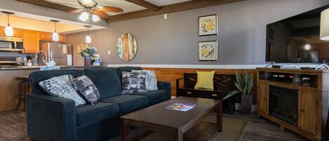 A living room with a sofa, coffee table, and a TV. There is a kitchen area with wooden cabinets and a refrigerator.