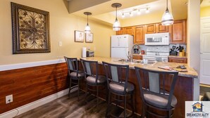 A kitchen with wooden cabinets, a refrigerator, a microwave, and a bar area with bar stools. - Walk-In Branson Condo w/ Pool + Easy Access | Club C-7 (Branson)