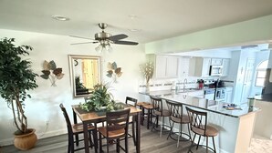 Una cucina moderna con isola centrale, tavolo da pranzo in legno e sedie, una ventola a soffitto e piante in vaso.