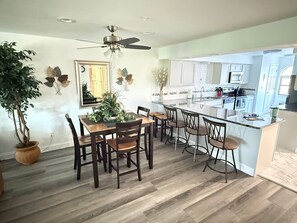 Eine moderne Küche mit einer zentralen Kochinsel, einem Holzespeisetisch und Stühlen, einem Deckenventilator und Topfpflanzen.