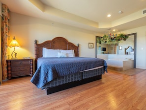A bedroom with a wooden bed, a nightstand, a lamp, a dresser, a large mirror, and a bathroom with a bathtub.