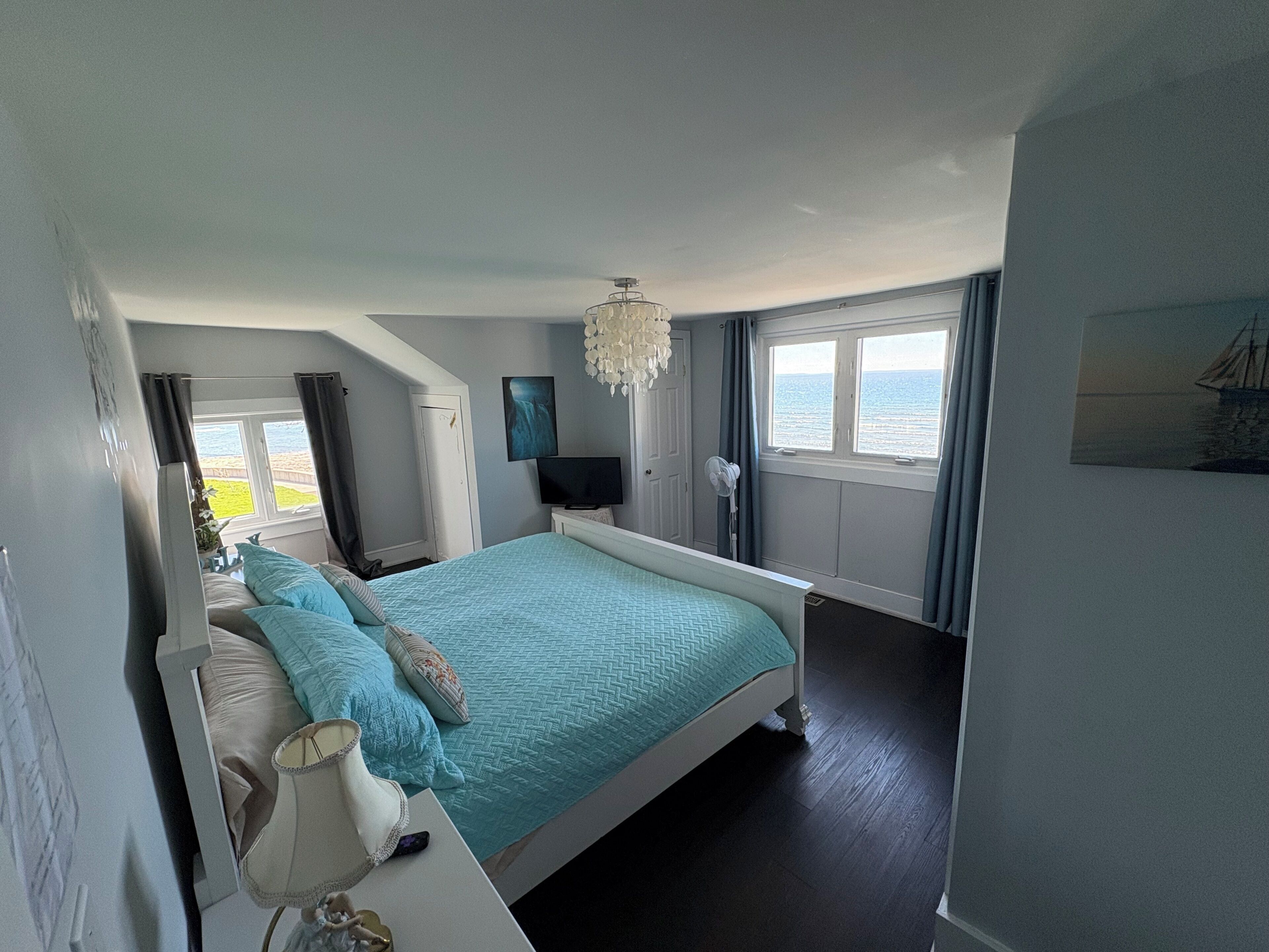 A bedroom with a bed, a TV, a chandelier, a window with a view of the sea, and a painting on the wall.
