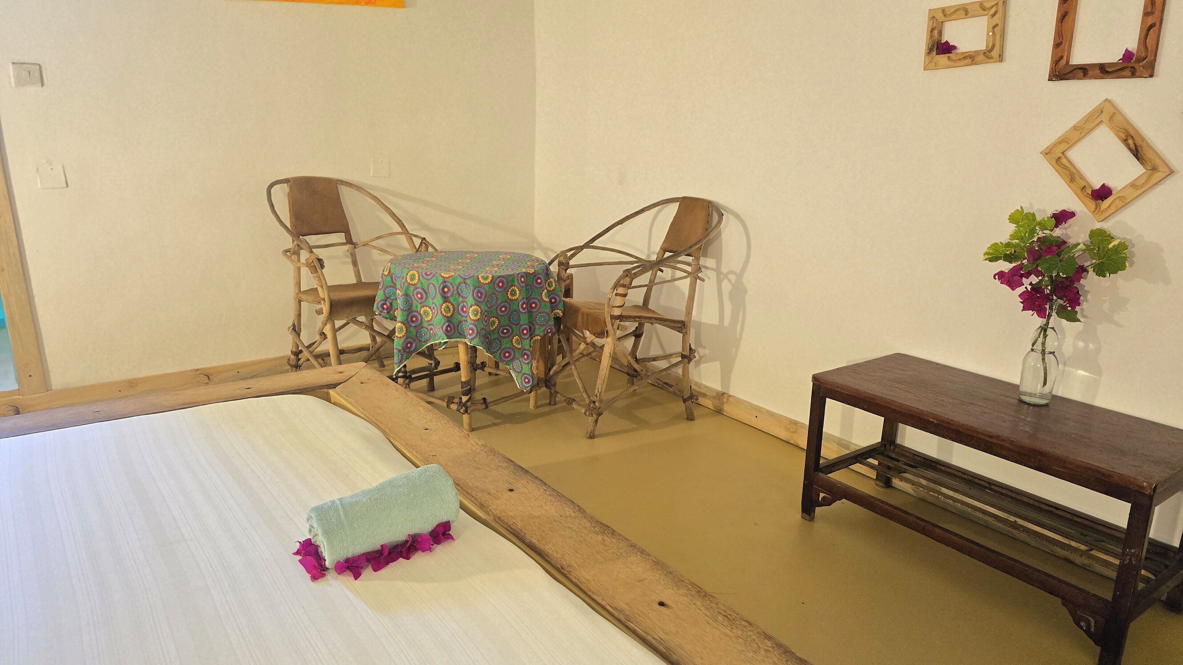 Une pièce comprenant un lit, deux chaises en bois, une petite table recouverte d’une nappe et un vase de fleurs posé sur une étagère en bois.