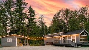 A cabin with a porch and string lights, surrounded by tall trees and a sunset sky. - Eureka Tiny Home w/ Hot Tub & Community Beach! (Eureka)