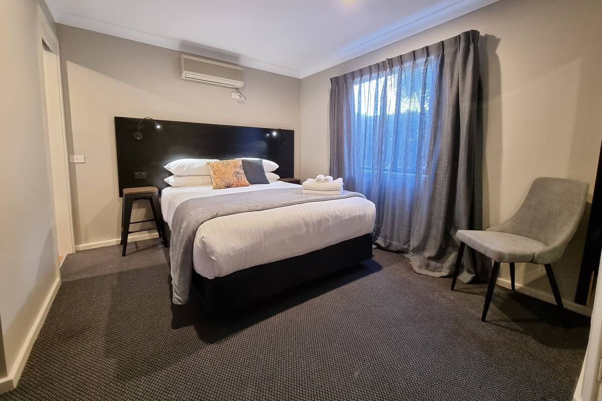 Une chambre d’hôtel avec un grand lit, une table de chevet, une chaise et une fenêtre avec des rideaux.
