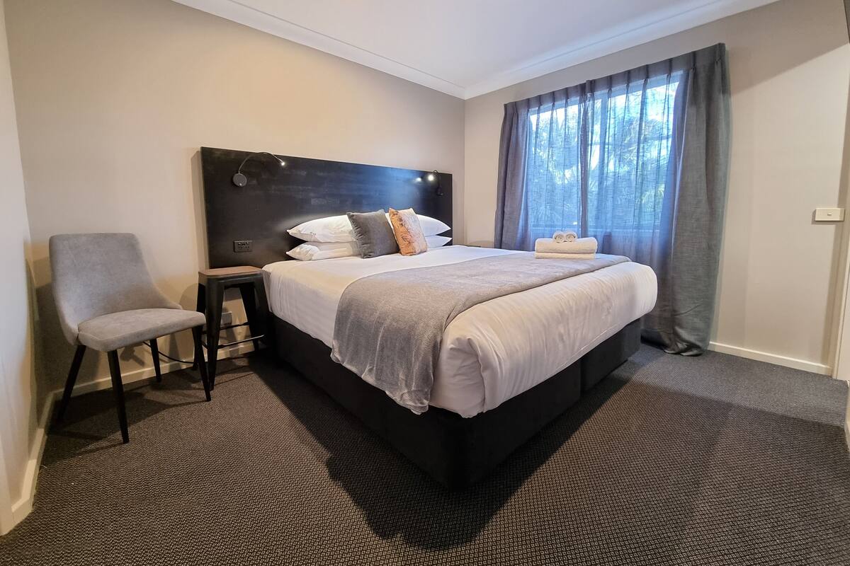 Une chambre d’hôtel avec un grand lit, une chaise, une table de chevet et une fenêtre avec des rideaux.