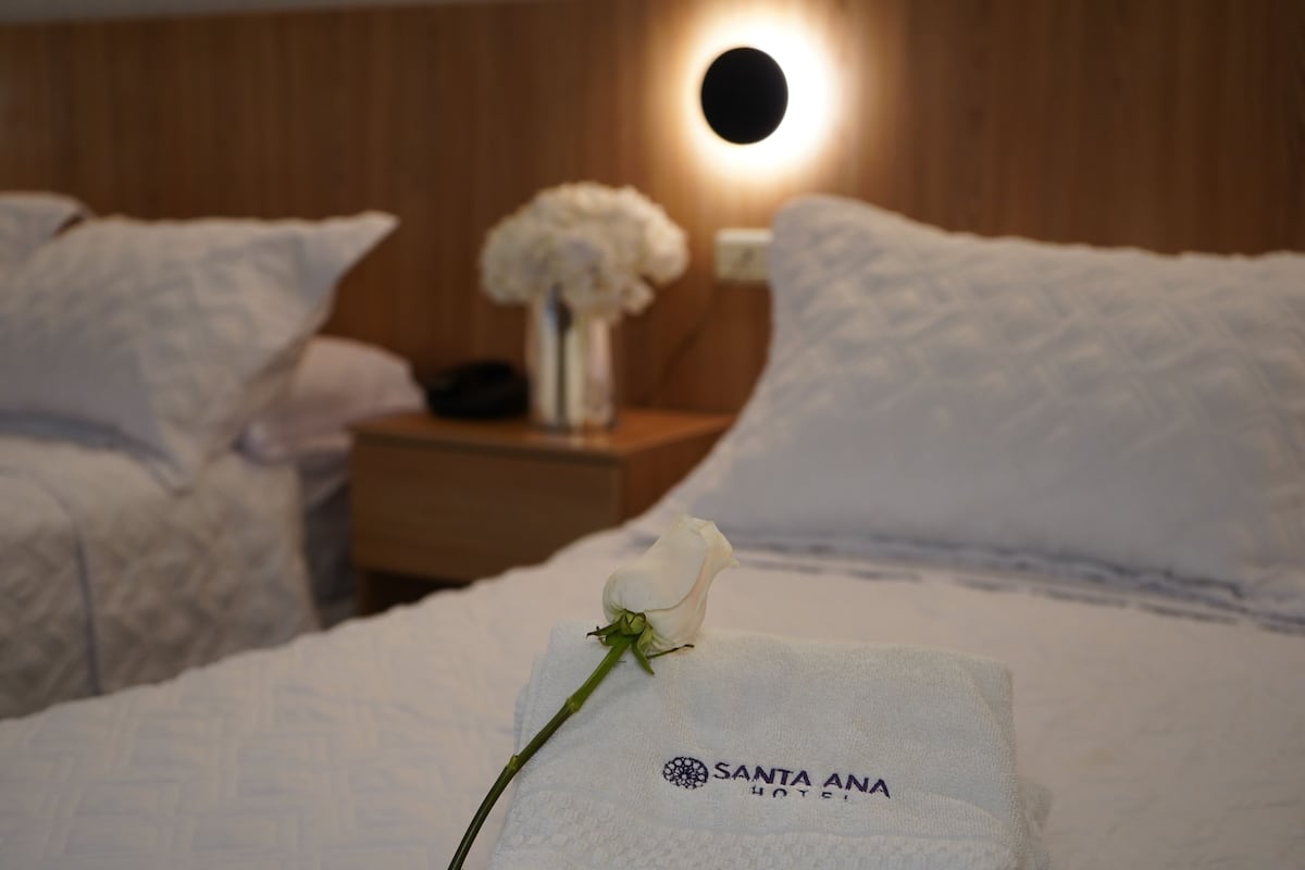 Ein Hotelzimmer mit einem ordentlich bezogenen Bett, einer weißen Rose auf dem Kopfkissen und einem Nachttisch mit Blumen in einer Vase.