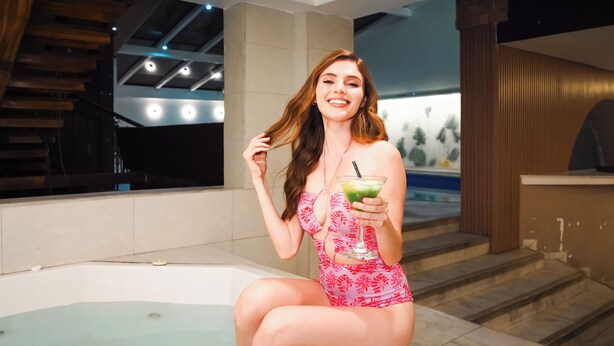 Una mujer con traje de baño rosa sentada junto a una bañera en una habitación moderna con vista a una piscina y una escalera de madera.