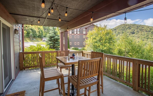 A wooden deck with a table and chairs, overlooking a forested area with a building in the background.