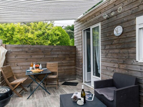 A covered patio area with a table set for dining, wooden chairs, a wicker sofa, and a bottle of wine.
