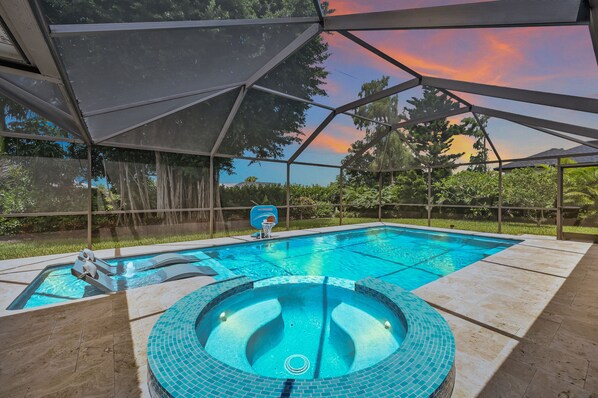 A swimming pool with a hot tub, surrounded by a covered area with a view of trees and a sunset. - Beautiful Naples Retreat  (Naples)