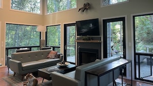 A spacious living room with a large sofa, a coffee table, and a fireplace. There are multiple windows providing views of the surrounding trees. - Private Wooded Acre, Soaring Windows in Nat’l Park, 2 blocks to the beach (Gary)