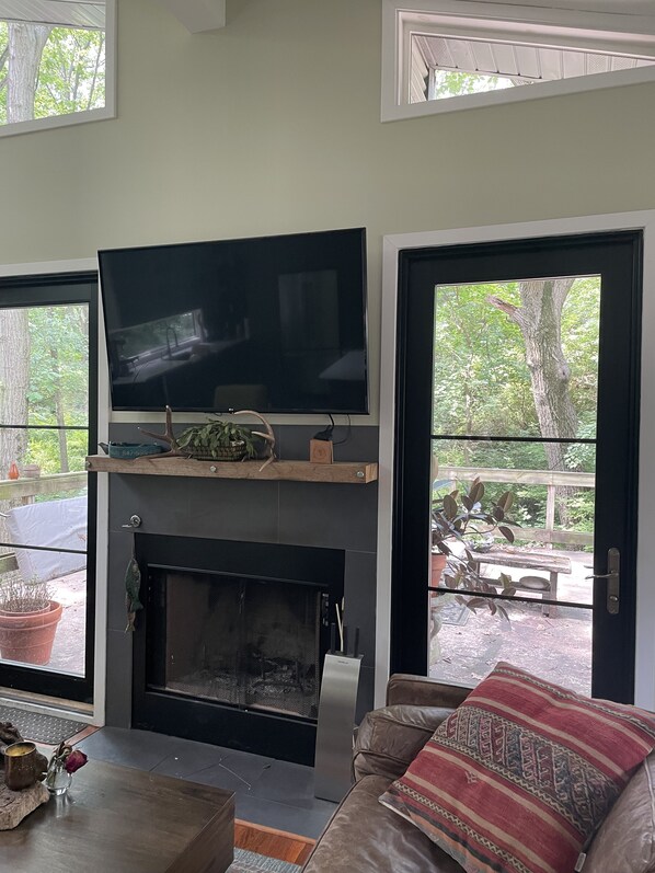 A living room with a flat-screen TV mounted above a fireplace, a sofa with a patterned cushion, and a view of a wooden deck and trees through large windows.