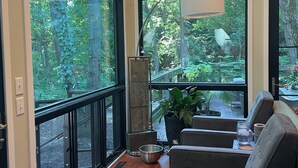 A spacious room with large windows offering a view of a lush garden. There is a comfortable seating area with a sofa and a side table. - Private Wooded Acre, Soaring Windows in Nat’l Park, 2 blocks to the beach (Gary)