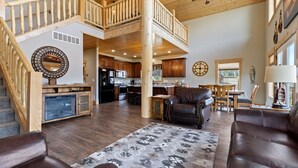 A spacious living area with a high ceiling, wooden staircase, a kitchen with dark wood cabinets, and a dining area with a view of the outdoors. - The Yellowstone Hideaway - Beautiful 3 Bedroom, 2 Bath Cabin- Sleeps 10 (Island Park)