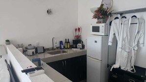 A compact kitchen with a refrigerator, microwave, sink, and a small countertop. There are white robes hanging on a rack, and a trash bin is visible.