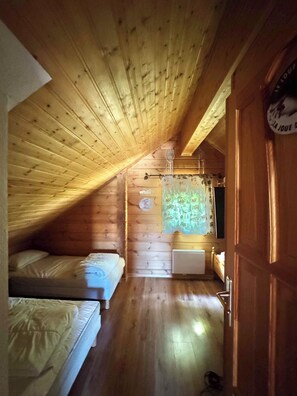 A room with wooden walls and a sloped ceiling, featuring two beds, a radiator, and a window with a curtain.