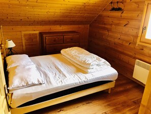 Quarto de cabana de madeira com uma cama, mesa de cabeceira, cômoda e janela.