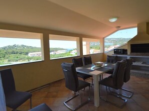 Uma área de refeições com mesa e cadeiras, uma cozinha com micro-ondas e grandes janelas com vista para uma encosta.
