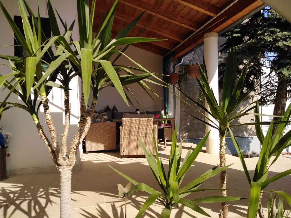 Uma área externa coberta com plantas, um sofá de vime e uma mesa com objetos sobre ela.