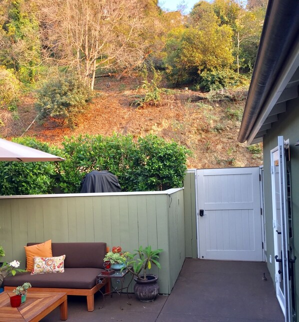 A patio area with a wooden bench, a small table, and a grill. There is a green fence and a white gate.