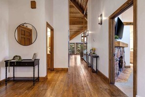 A hallway with wooden flooring, a round mirror, a wooden door, and a television mounted on a stone wall.