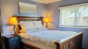 Un dormitorio con una cama de madera, dos mesitas de noche con lámparas, una ventana con persianas y un cuadro en la pared.