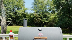 Un barbecue au charbon de bois sur une terrasse extérieure dotée d’une rambarde blanche, une bouteille d’allume-feu pour barbecue et un sac rouge.