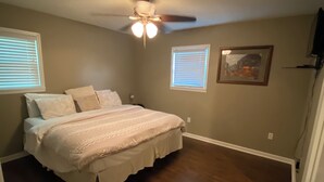 Une chambre à coucher avec un lit, deux fenêtres, un ventilateur de plafond et un tableau accroché au mur.