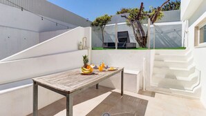 A modern outdoor patio with a wooden table set for two, a small tree, and a white wall.